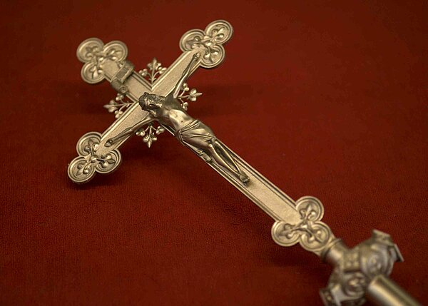 Bild: Ein kleines Goldenes Kreuz, welches auf einem rotem Stück Stoff liegt.
