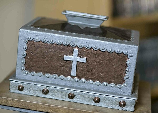 Bild: Ein kleines braun silbernes Kästchen mit einem Kreuz darauf.