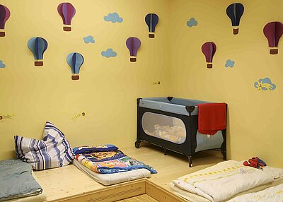 Viele Matratzen und ein Beistellbett im Kinderhaus Kunterbunt in Grafenwöhr. An der Gelben Wand des Zimmers hängen selbstgebastelte Heißluftballons und Wolken aus Papier