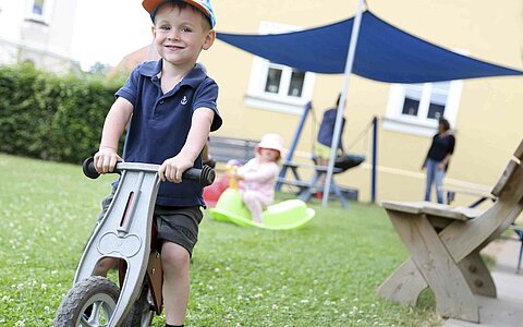 Ein kleiner Junge aus dem Haus für Kinder Gottfried Sperl in Vohenstrauß, der auf einem grauen Laufrad sitzt. Im Hintergrund sieht man ein weiteres Kind auf einer Wippe, eine Sitzbank und ein Sonnensegel.