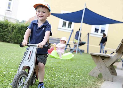 Ein kleiner Junge aus dem Haus für Kinder Gottfried Sperl in Vohenstrauß, der auf einem grauen Laufrad sitzt. Im Hintergrund sieht man ein weiteres Kind auf einer Wippe, eine Sitzbank und ein Sonnensegel.