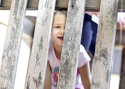 Ein Mädchen, welches durch die Holzstäbe des Spielhauses aus dem Garten der St. Michael Kita in Weiden schaut.