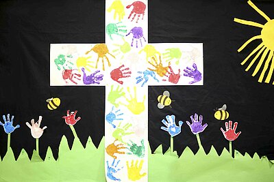 Ein Bild von einem weißen Jesuskreuz, auf dem ganz viele kleine bunte Kinderhände darauf sind. Darunter befindet sich eine Wiese, bei der die kleinen Hände der Kinder als Blumen dargestellt wurden