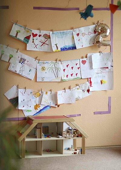 Eine Orange Wand in dem Haus für Kinder Gottfried Sperl in Vohenstrauß, welche mit selbst gemalten Bildern von den Kindern vollhängt. Unter den Gemälden steht ein kleines Puppenhaus
