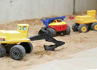Ein gelber Spielzeugbagger in einem Sandkasten. Im Hintergrund befinden sich ein Spielzeugkipper und ein weiterer Spielzeugbagger.