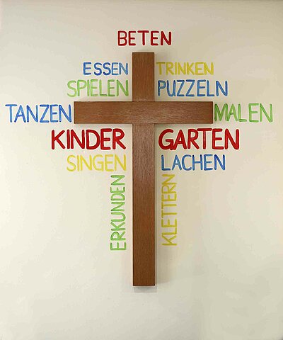 Ein Jesuskreuz an der Wand der St. Michael Kita in Weiden. Außen um das Kreuz stehen viele bunte Wörter, mit Dingen, die man im Kindergarten machen kann.