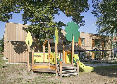Ein kleines Spielehaus mit integrierter Rutsche im Garten der Kinderscheune Kreuz Christi in Weiden. Im Hintergrund ist der Kindergarten zu sehen.