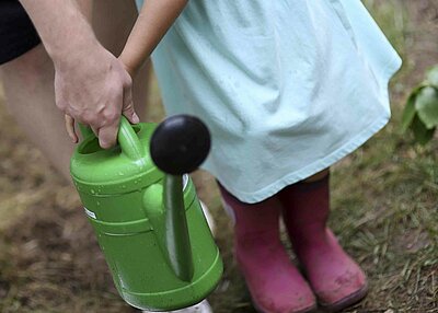 Ein kleines Mädchen aus dem St. Markus Kindergarten in Weiden, welche mit Hilfe eines Erziehers eine grünen Gießkanne in der Hand hält-