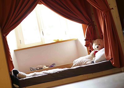Ein Bett in dem evangelischen Kindergarten Regenbogen in Erbendorf mit einem Teddybären und Roten Gardienen darüber.
